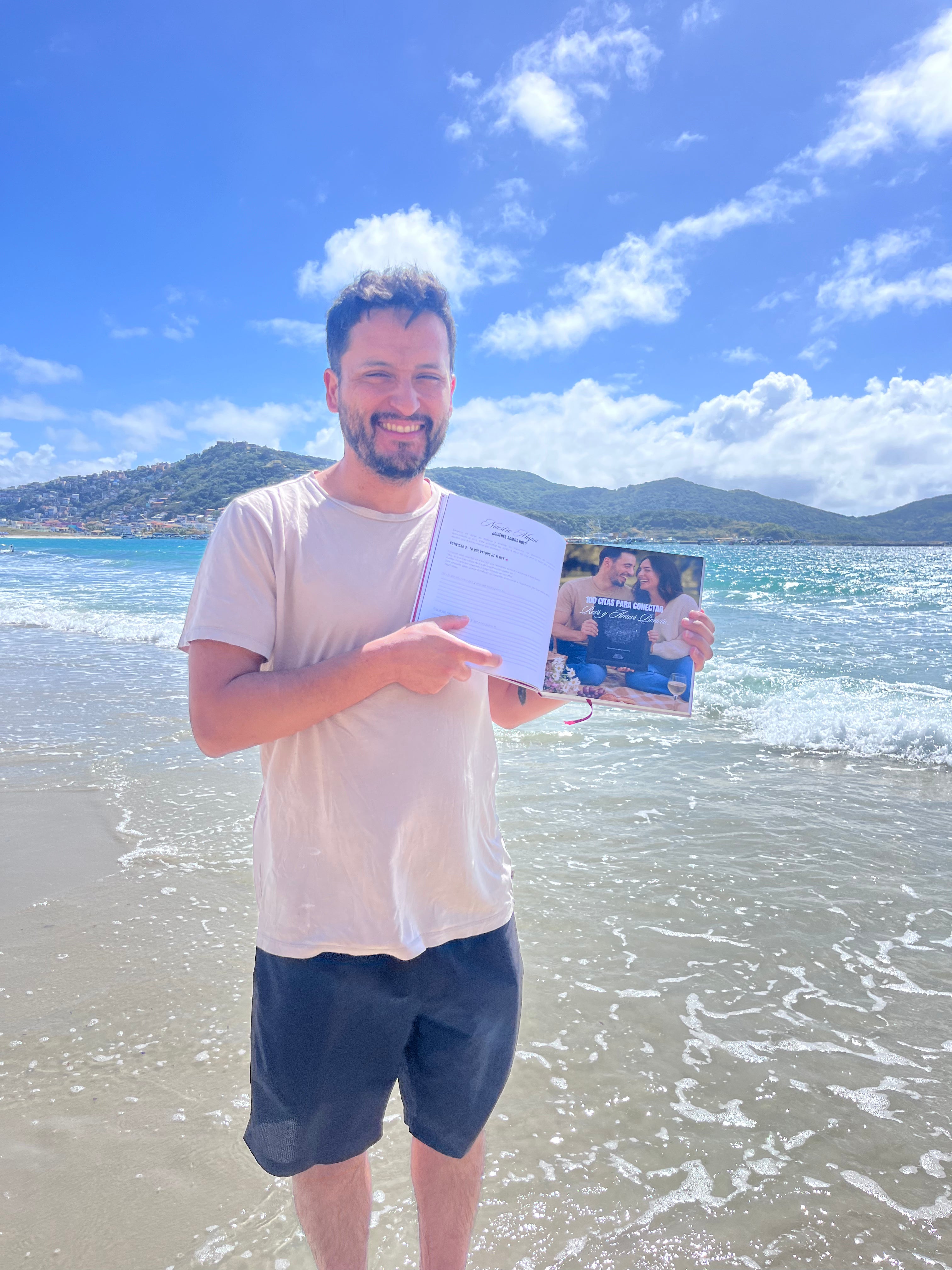 Hombre en la orilla de la playa, sonriendo y señalando el Álbum para Parejas “Esto va en serio” abierto, mostrando una página con foto de pareja y texto, ideal para registrar citas y recuerdos juntos.