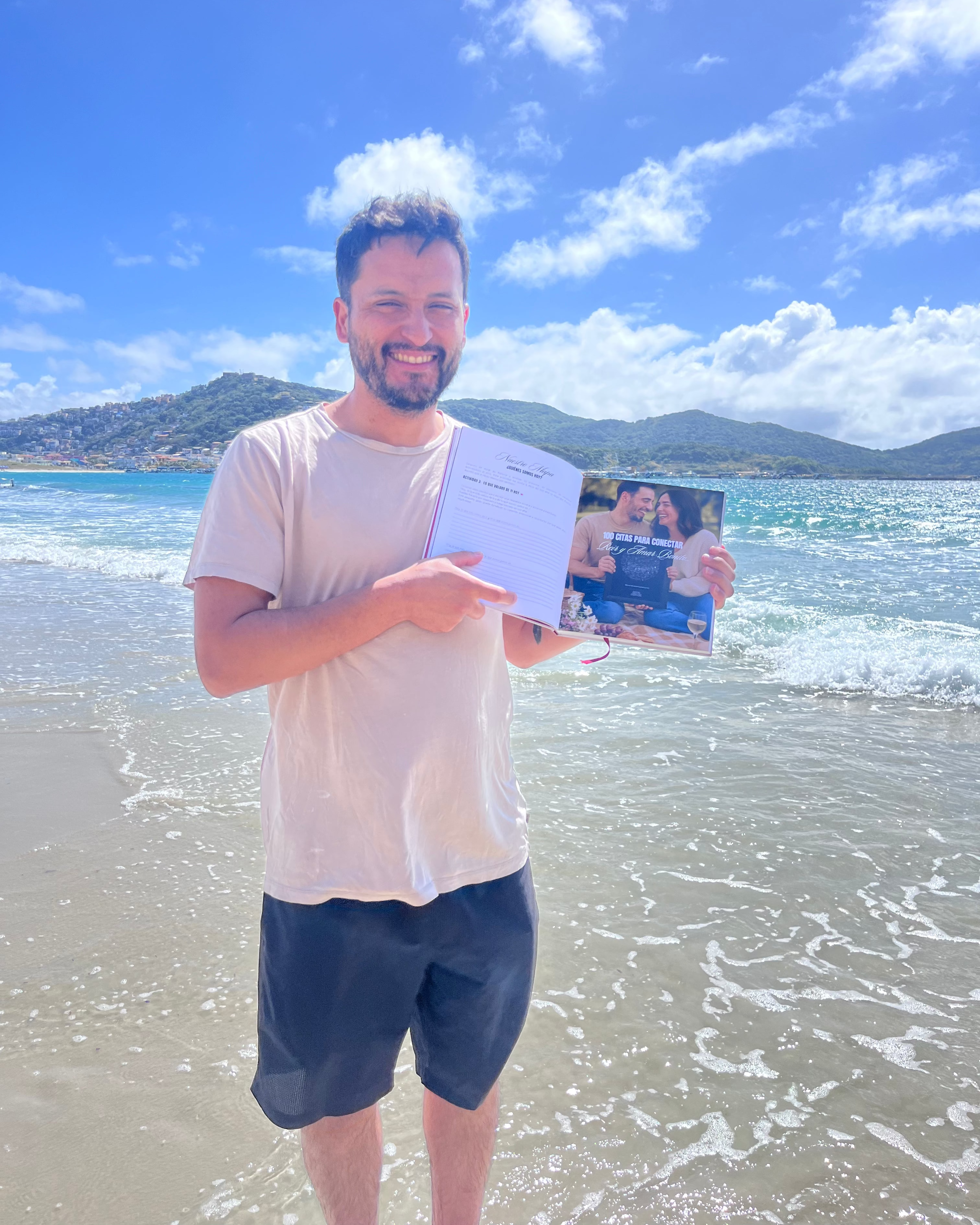 Hombre en la orilla de la playa, sonriendo y señalando el Álbum para Parejas “Esto va en serio” abierto, mostrando una página con foto de pareja y texto, ideal para registrar citas y recuerdos juntos.