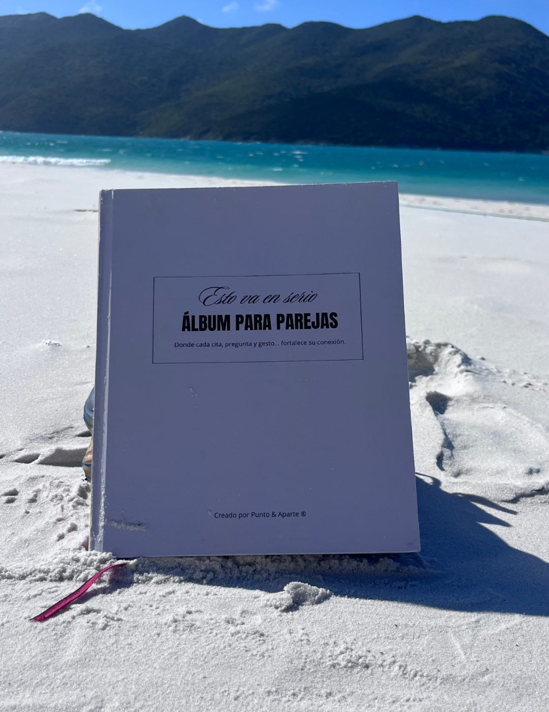 Álbum para parejas “Esto va en serio” sobre la arena blanca de la playa, con mar turquesa y montañas de fondo.