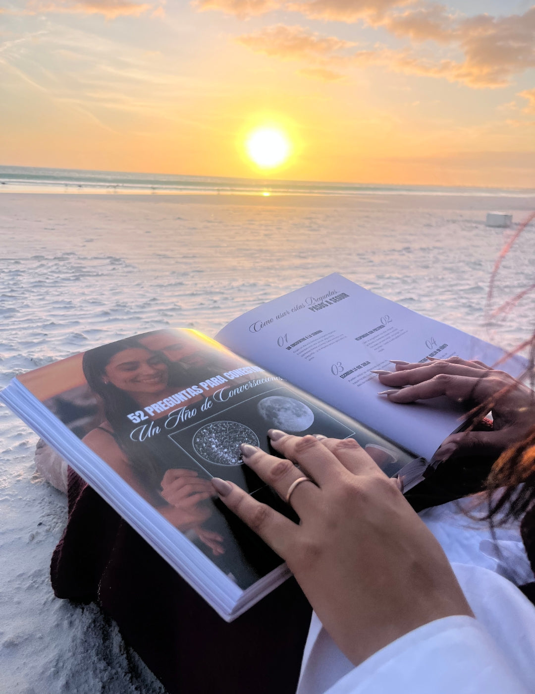 Mujer leyendo el Álbum para Parejas “Esto va en Serio” sobre la arena al atardecer, con el mar de fondo, mostrando una cita romántica al aire libre.
