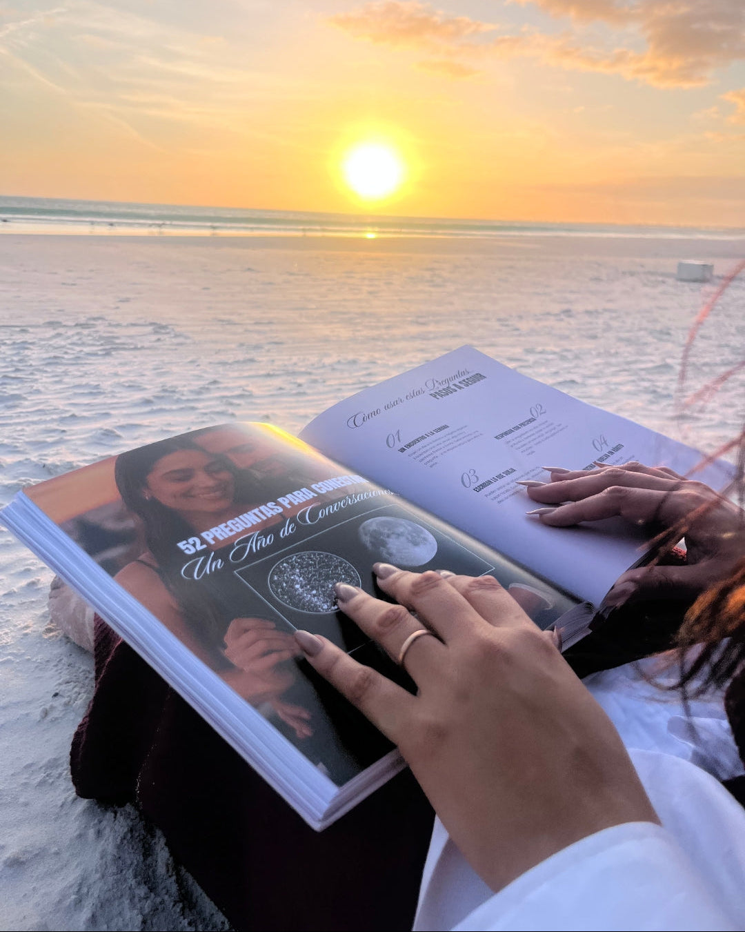 Mujer leyendo el Álbum para Parejas “Esto va en Serio” sobre la arena al atardecer, con el mar de fondo, mostrando una cita romántica al aire libre.