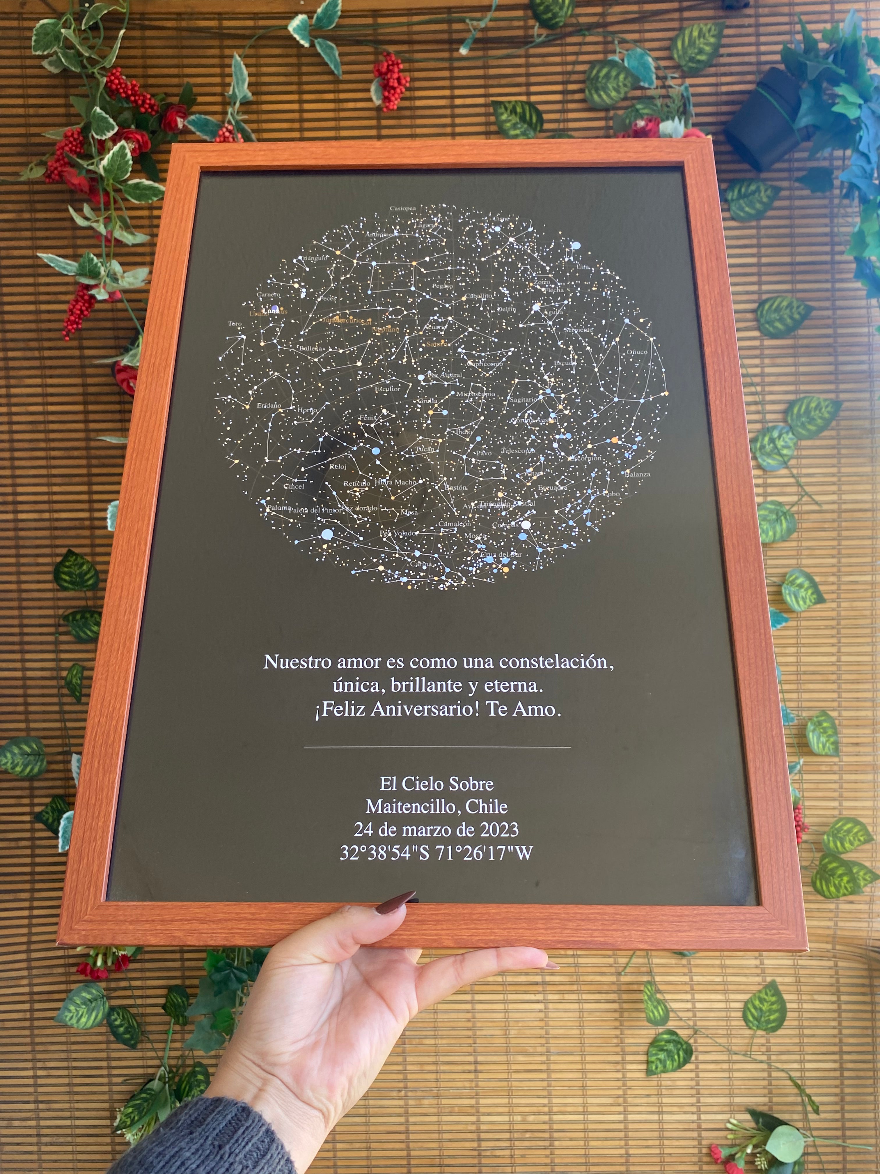 Mapa estelar XL con marco de madera sostenido por una mano, sobre fondo de bambú con hojas verdes, como regalo personalizado de aniversario.