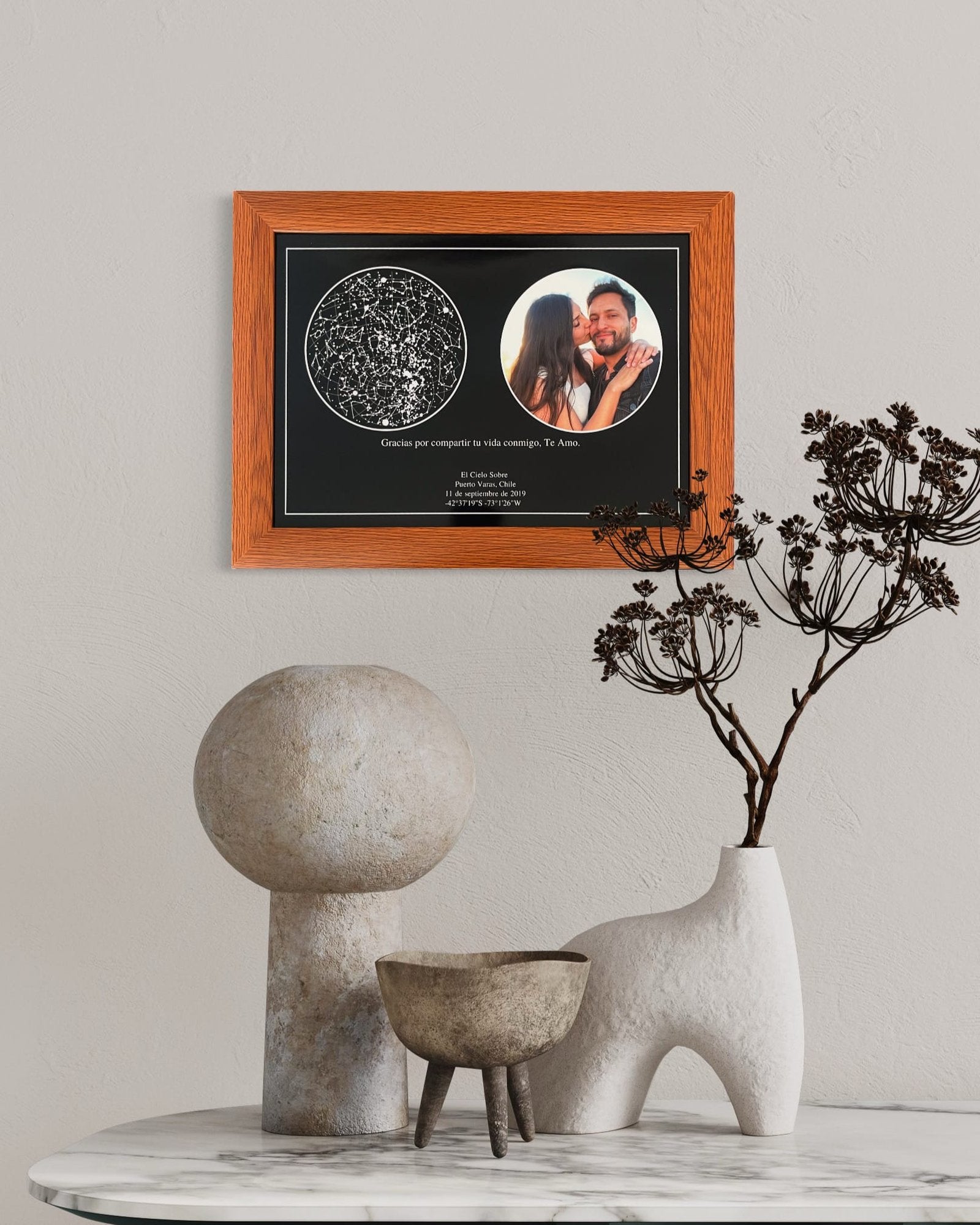 Mapa estelar con foto de pareja en marco de madera color miel, colgado sobre mesa decorativa con jarrones y esculturas modernas.