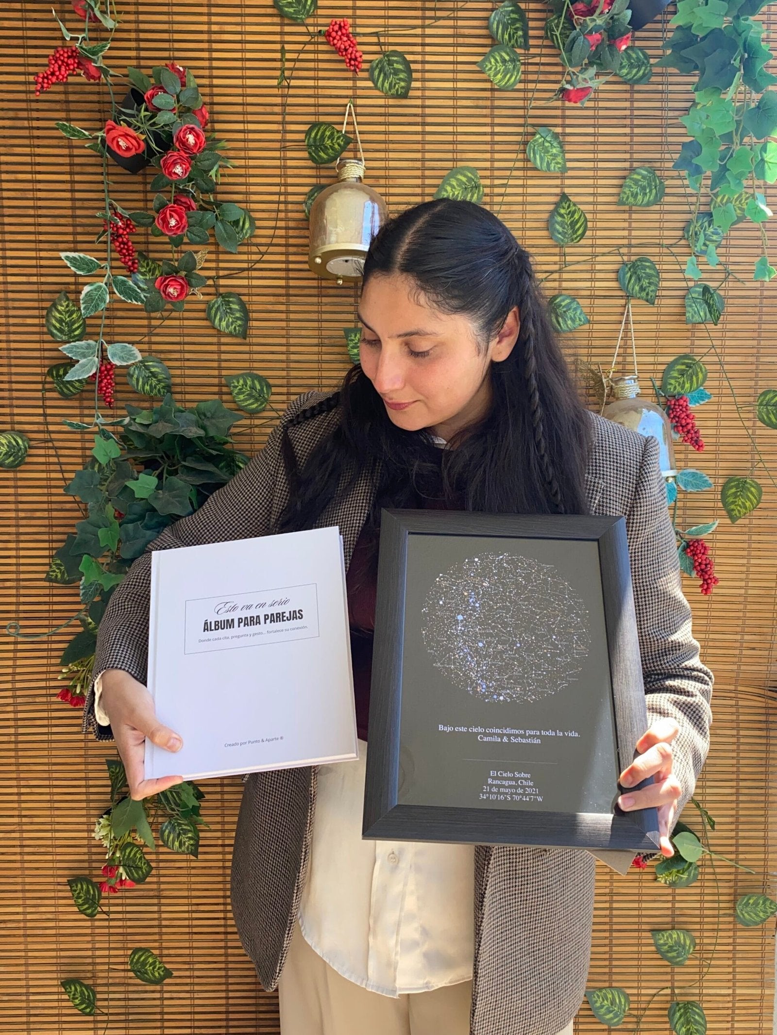 Mujer de pie frente a un fondo con plantas sosteniendo el Álbum para Parejas "Esto va en serio" y un Mapa Estelar Sky of Colors en marco negro.