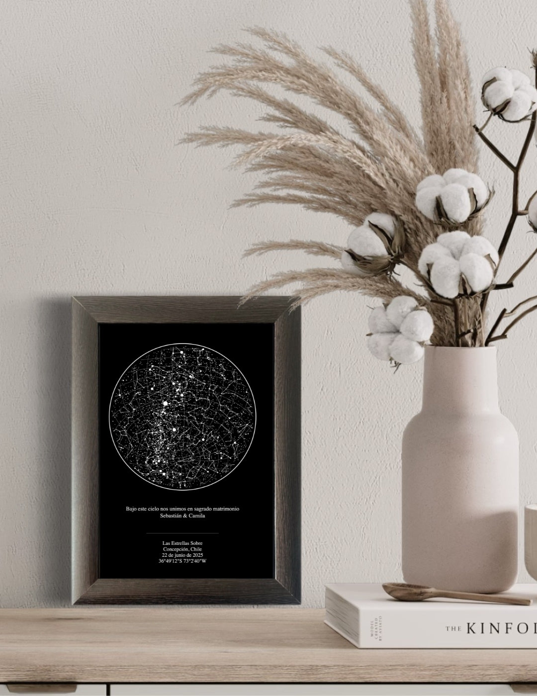 Mapa estelar tradicional enmarcado en color gris sobre una repisa de madera clara, acompañado de un florero rosa con pampas y algodón, ideal como decoración minimalista y elegante para el hogar.