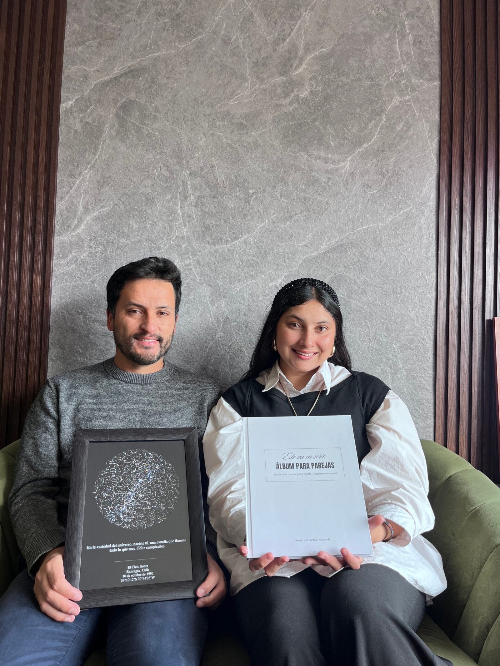 Pareja sentada en un sofá mostrando un Mapa Estelar Sky of Colors en marco negro y el Álbum para Parejas "Esto va en serio" en tapa dura.