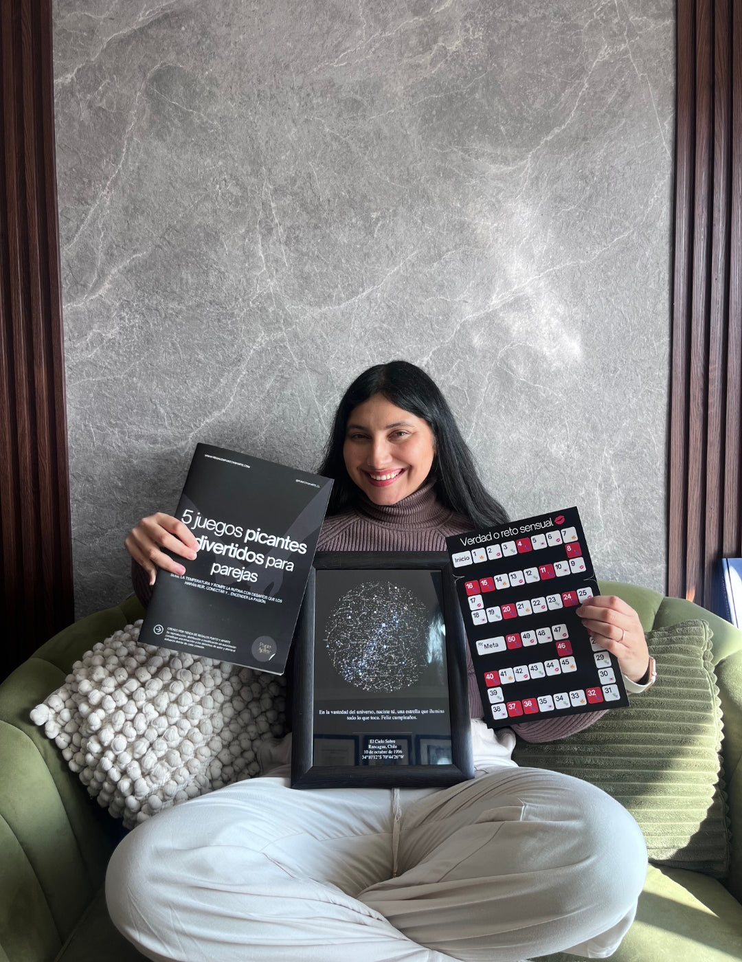 Mujer sentada en un sofá sosteniendo la revista 5 juegos picantes para parejas, el Mapa Estelar Sky of Colors y el tablero de juego del pack.