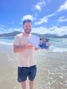Hombre en la orilla de la playa, sonriendo y señalando el Álbum para Parejas “Esto va en serio” abierto, mostrando una página con foto de pareja y texto, ideal para registrar citas y recuerdos juntos.