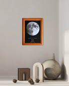 Mapa lunar enmarcado con borde de madera color miel colgado sobre una repisa decorativa con jarrón y figuras modernas.