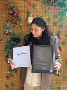 Mujer de pie frente a un fondo con plantas sosteniendo el Álbum para Parejas "Esto va en serio" y un Mapa Estelar Sky of Colors en marco negro.