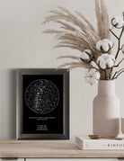 Mapa estelar tradicional enmarcado en color gris sobre una repisa de madera clara, acompañado de un florero rosa con pampas y algodón, ideal como decoración minimalista y elegante para el hogar.