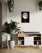 Cuadro Mapa Lunar XL enmarcado sobre un mueble de madera claro, decorado con planta grande y fotos familiares en un living acogedor.