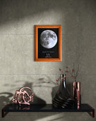 Cuadro con mapa lunar enmarcado en madera color miel sobre pared gris, decorando repisa moderna con jarrones y ramas secas. Ideal como regalo romántico y pieza de arte astronómica para el hogar.