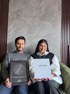 Pareja sentada en un sofá mostrando un Mapa Estelar Sky of Colors en marco negro y el Álbum para Parejas "Esto va en serio" en tapa dura.