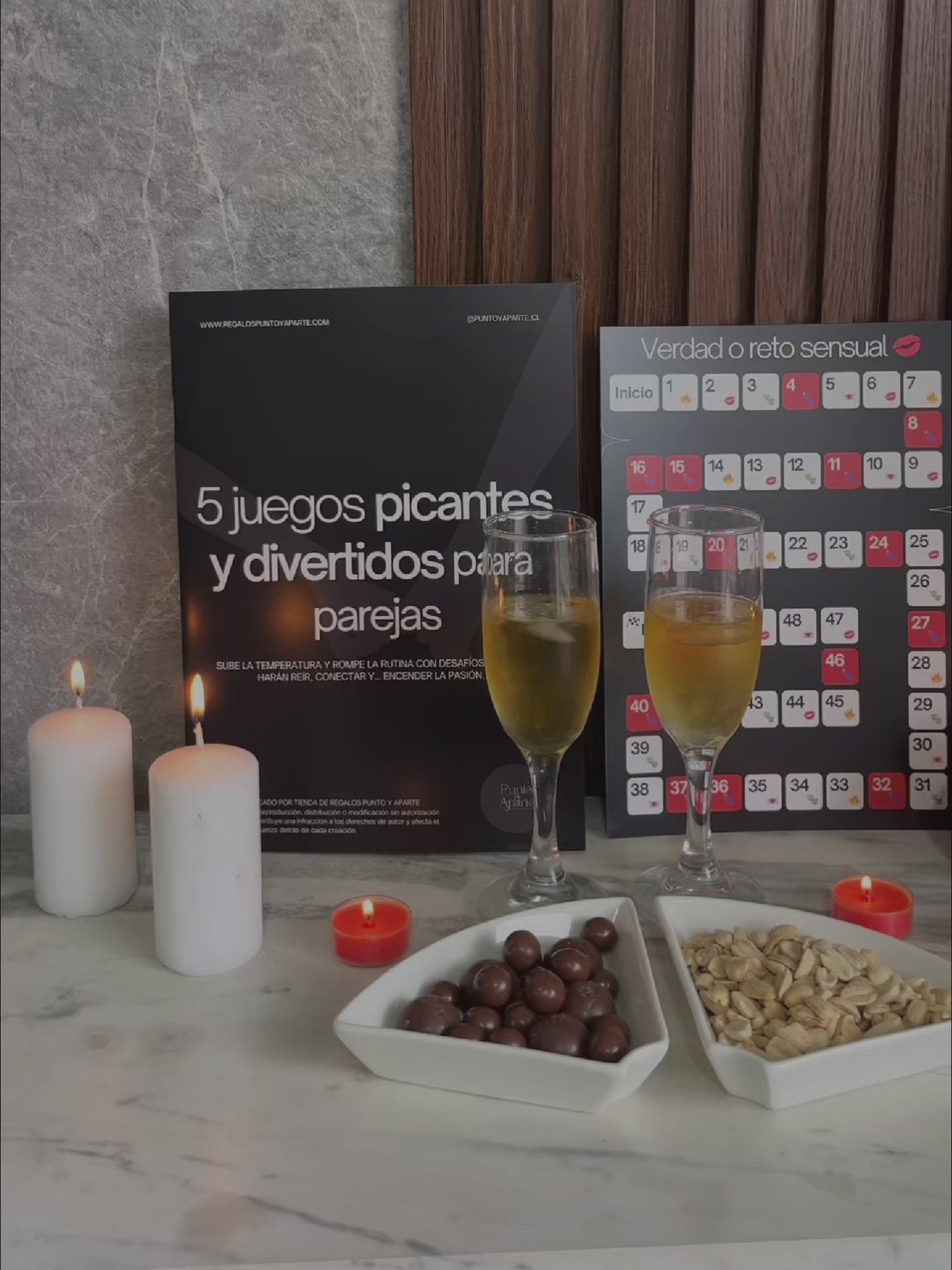 Video de la revista 5 juegos picantes para parejas junto a tablero del juego, copas de espumante, frutos secos y velas encendidas sobre una mesa.