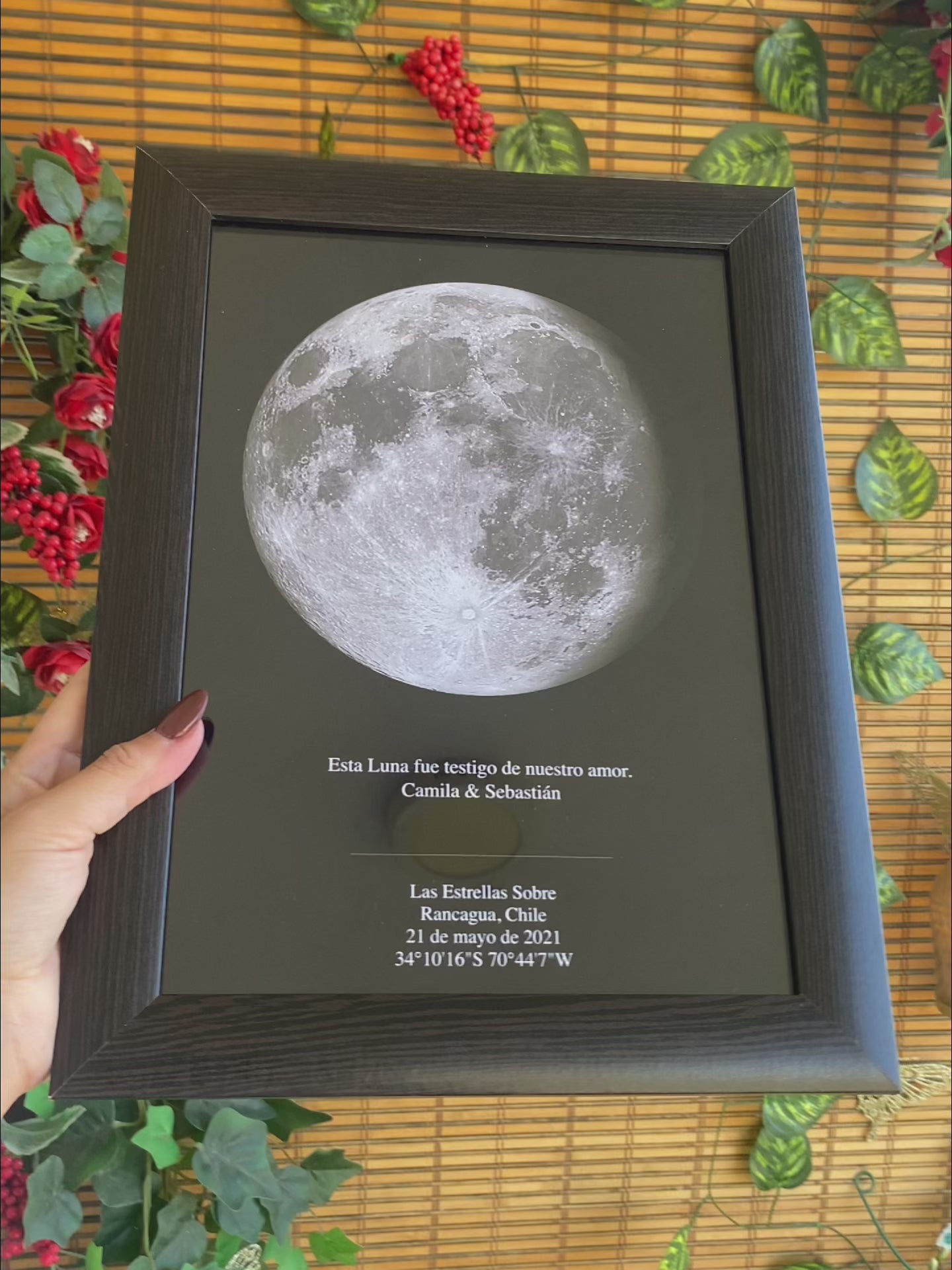 Mano sosteniendo un mapa lunar personalizado con frase romántica, en marco negro, sobre fondo de bambú con hojas verdes y detalles decorativos rojos.